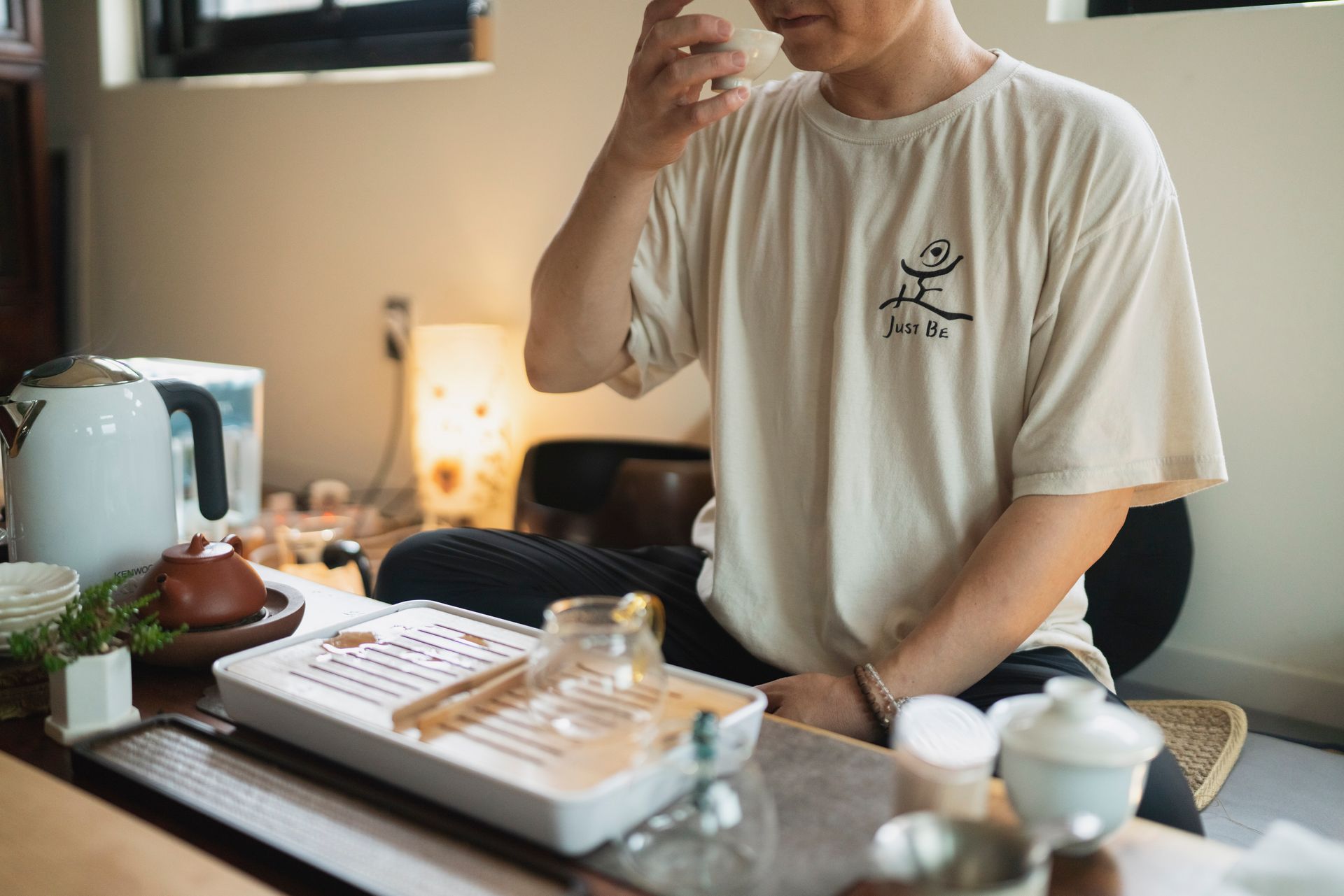Person sitting at a tea ceremony, pouring tea. Beige shirt, white tea tray, light-filled room.