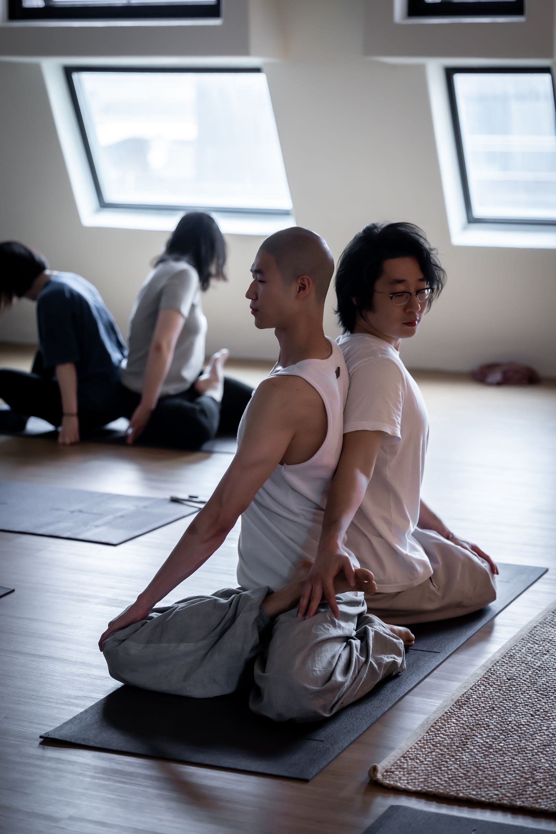 Two people in a yoga class seated back-to-back. Bald man in tank top, glasses wearing person, others in background.