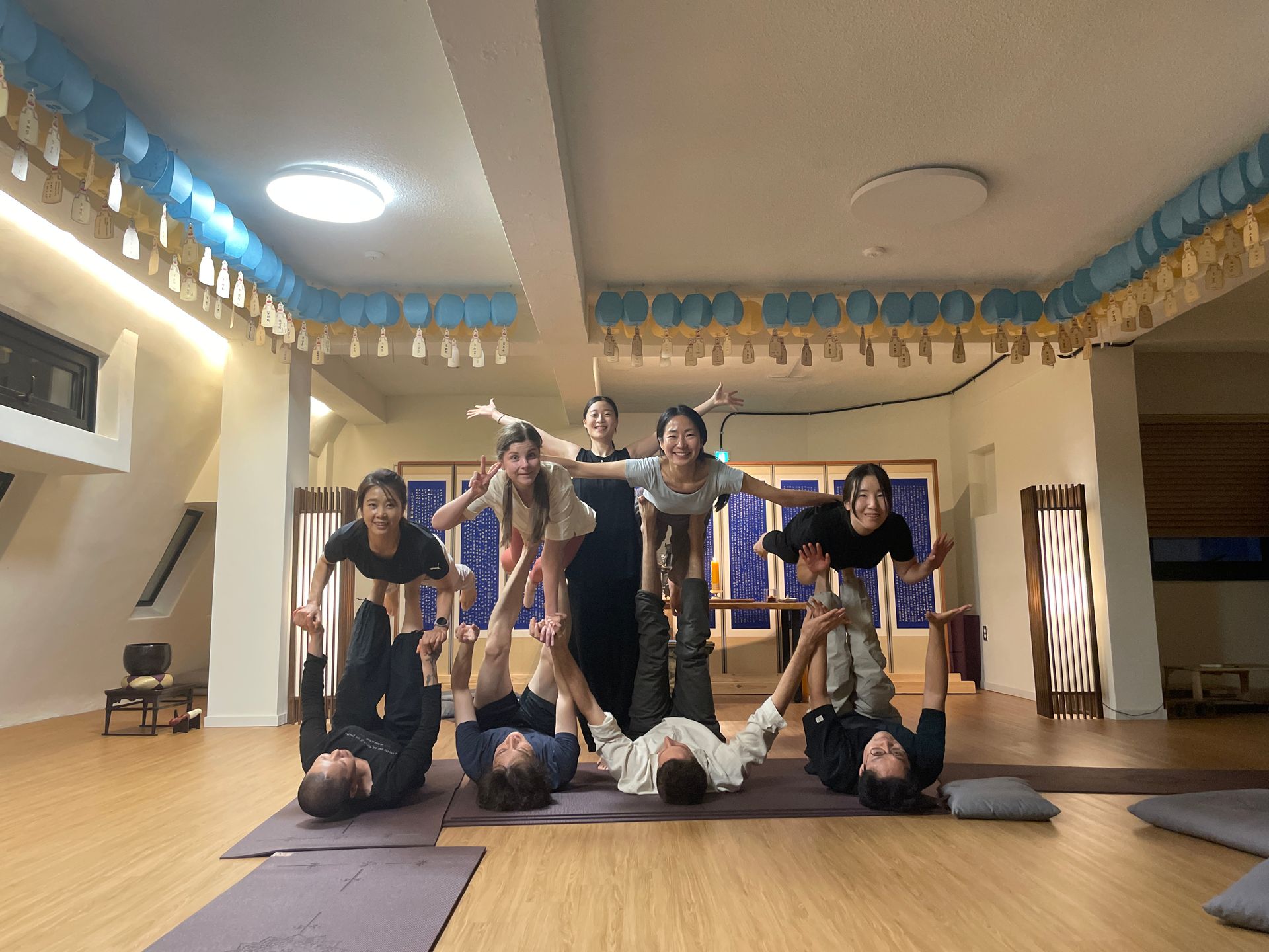 Group of people performing acro yoga indoors, arms outstretched, smiling.