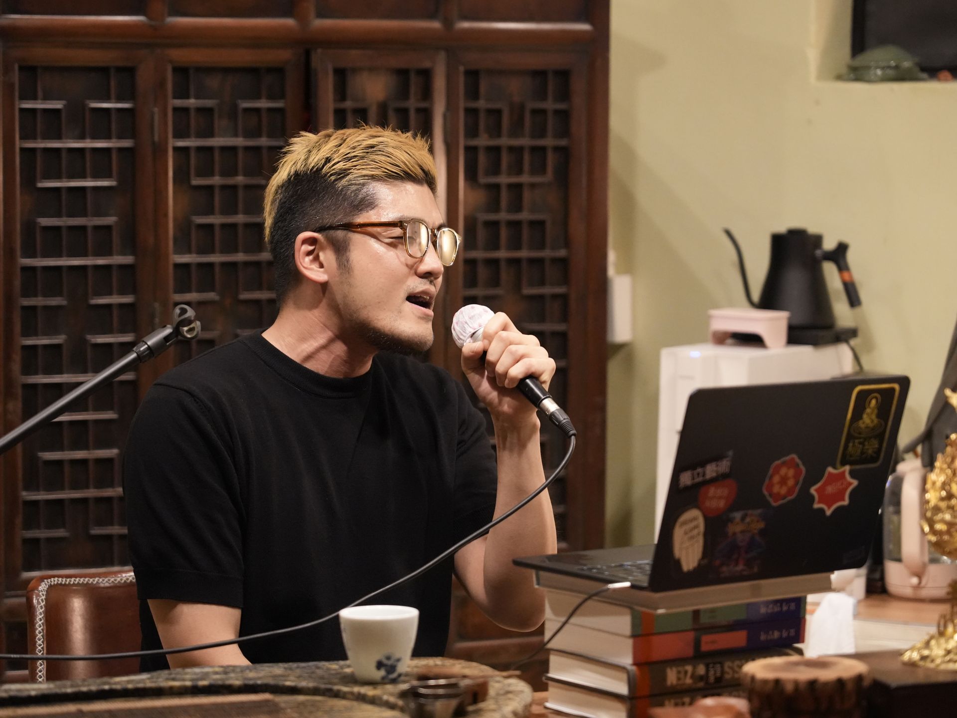 Man with blonde hair singing into a microphone, laptop, books, and tea set on table.