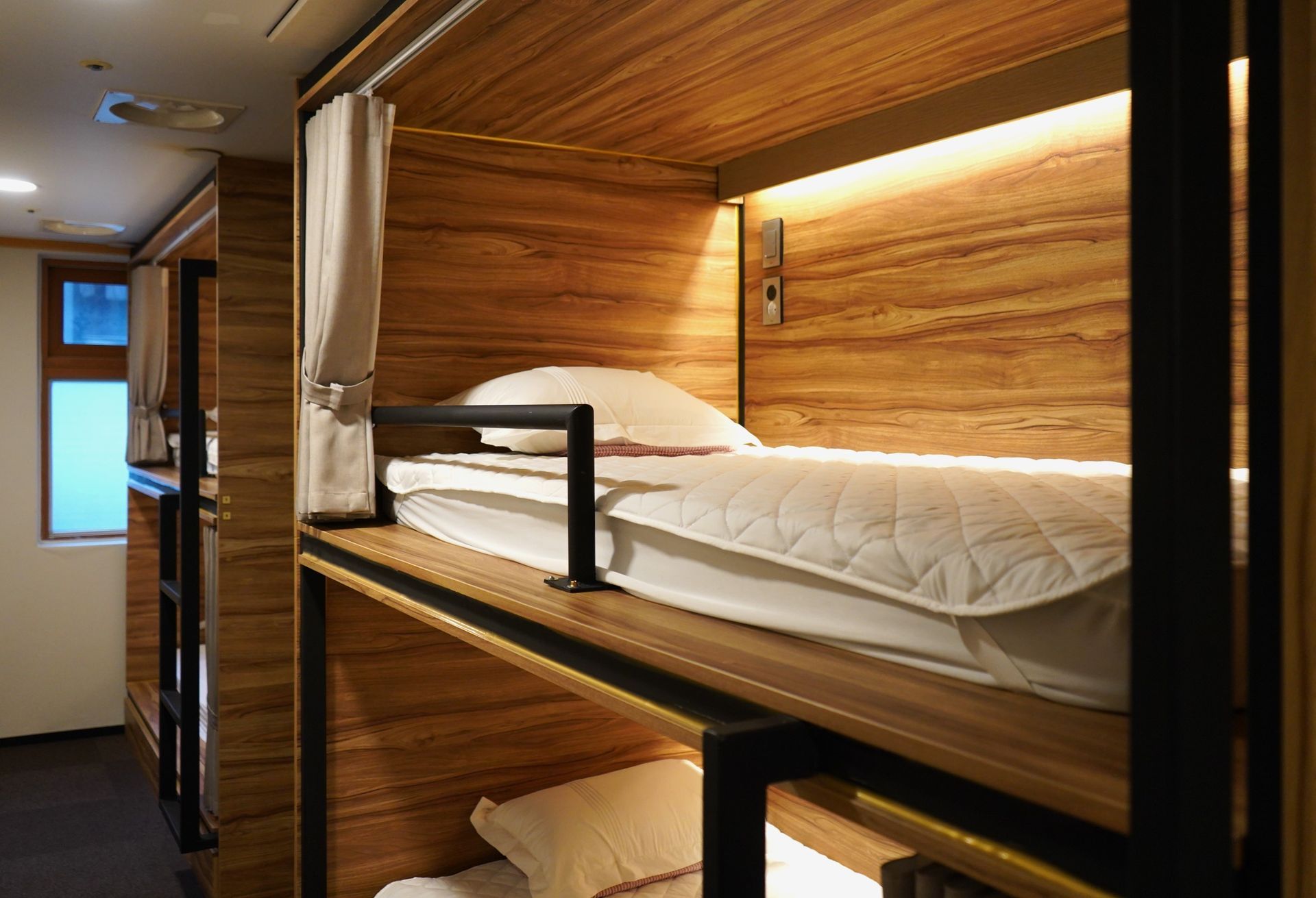 Wooden capsule hotel room with bunk beds, white bedding, and a small window.