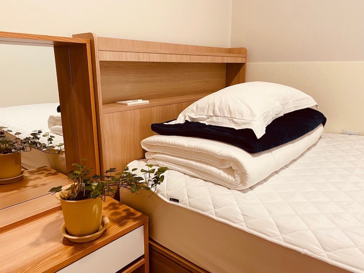 Cozy bedroom with a bed holding pillows and blankets. A potted plant sits on a nightstand with a mirror.