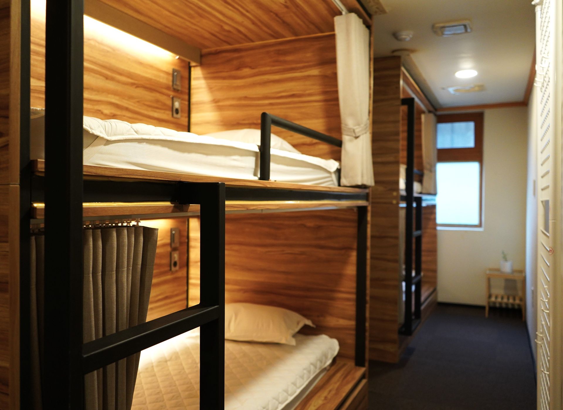 Bunk beds in a wooden capsule hotel room; a window with natural light is visible at the end of the hallway.