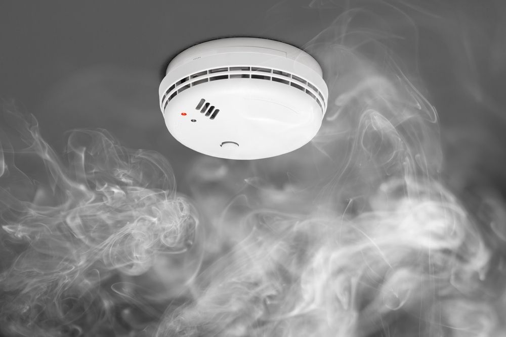 Smoke detector on a ceiling, surrounded by thick smoke, red light illuminated.