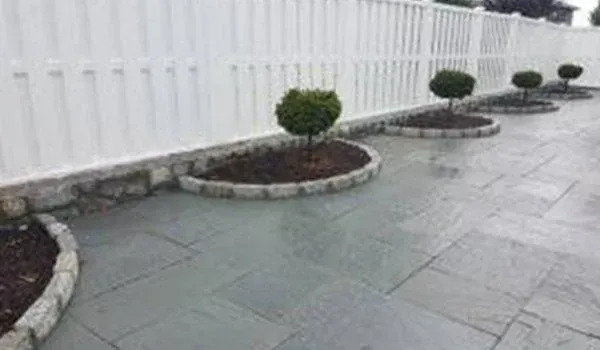 Gray stone patio with circular garden beds bordering a white fence; small trees in beds.