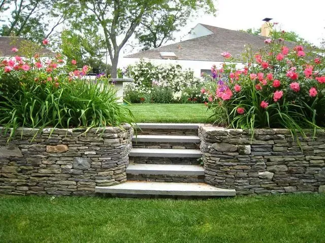 Stone steps lead up to a garden with pink flowers, a lawn, and a house in the background.