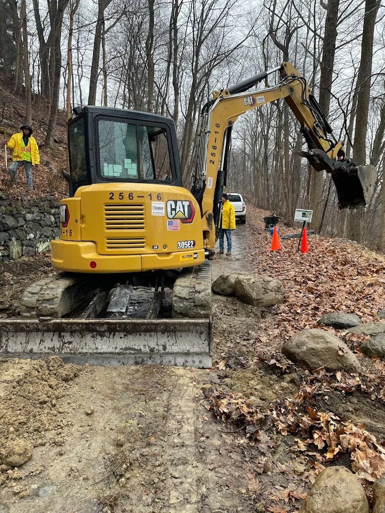 Yellow excavator on a muddy path, with workers in yellow vests; trees in the background.