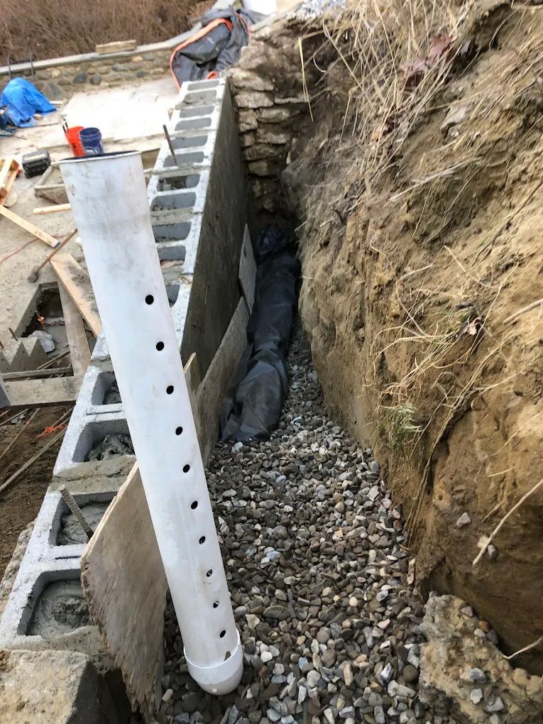 Construction of a retaining wall and drainage system. A perforated pipe runs through gravel, against a black membrane.
