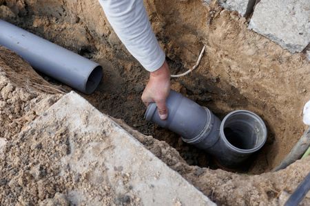 Person connecting gray plastic pipes in a trench, likely for drainage or plumbing.