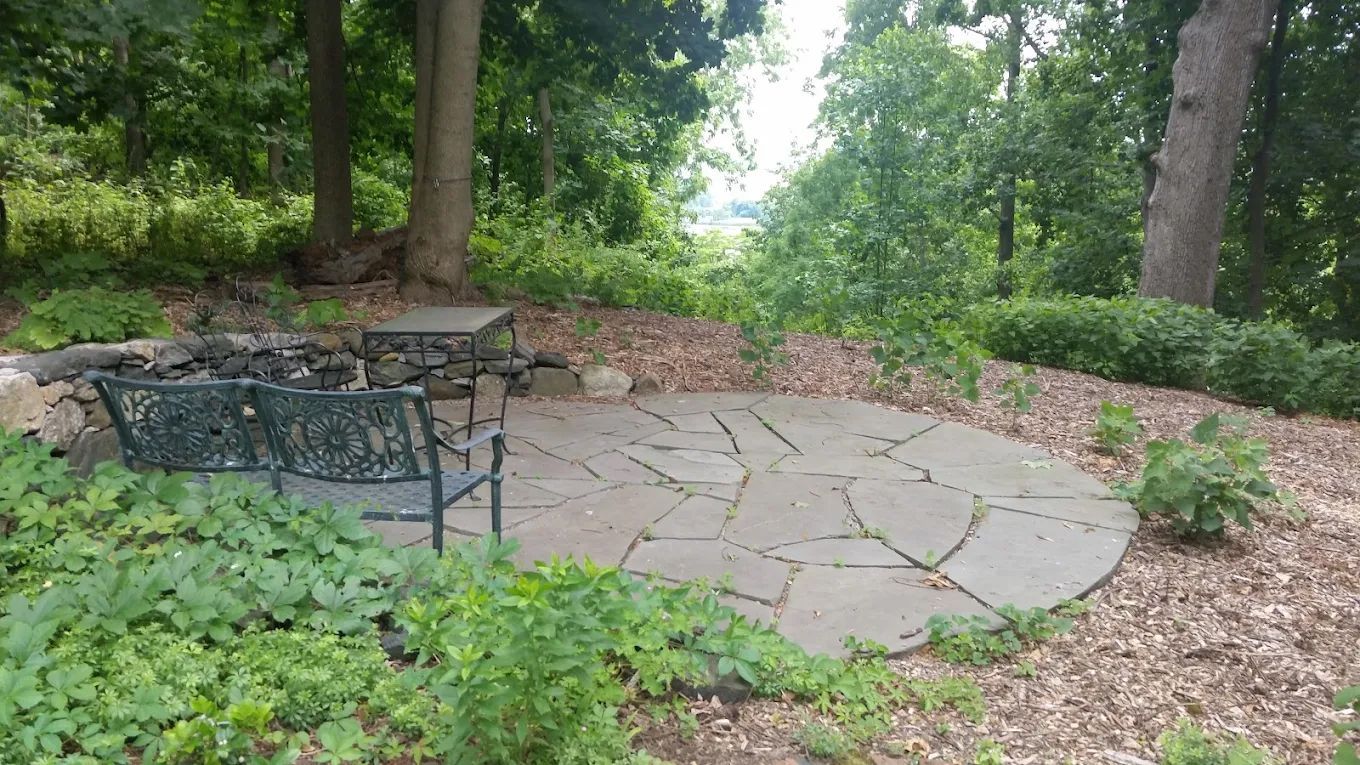 Stone patio with benches, fire pit, and surrounding greenery in a wooded area.