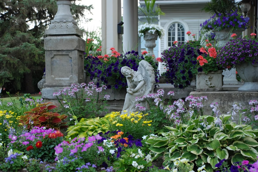 Garden bed with colorful flowers, stone angel statue, and urns in front of a house.