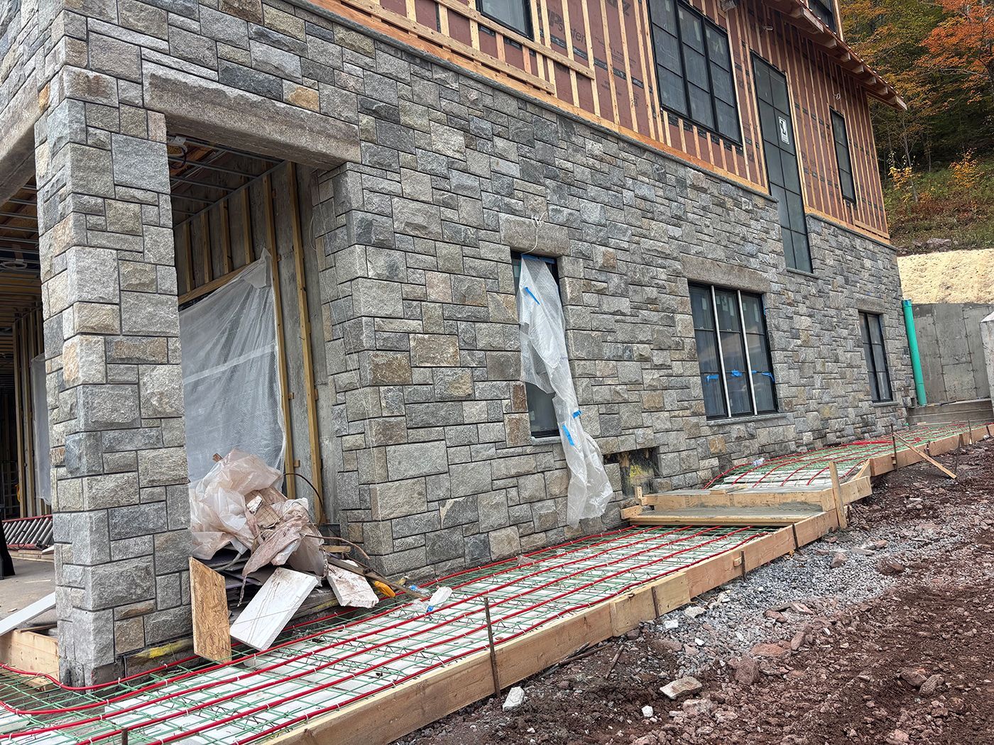 Stone-clad building under construction.  Exterior with windows, wood trim, and a concrete pathway with red tubes.