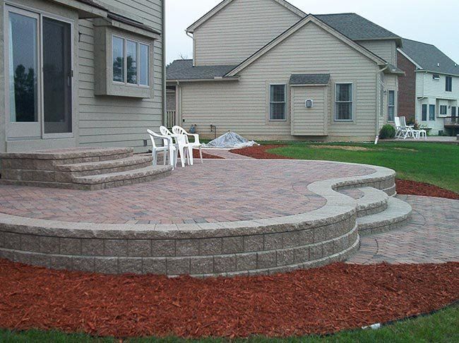 Brick patio with steps, retaining wall, and two houses in the background. Red mulch and white chairs.