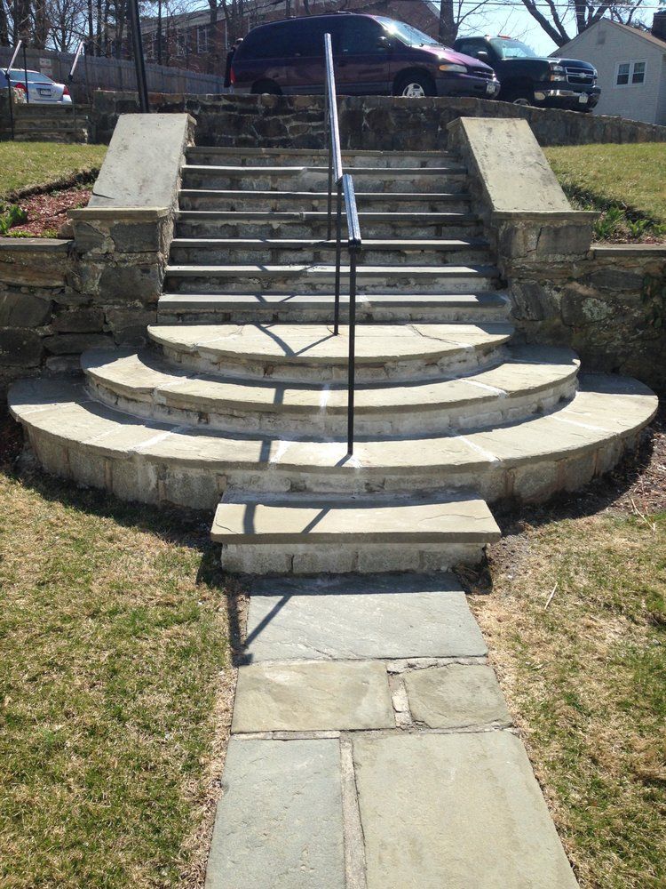 Stone steps leading up to a house, with a handrail. Pathway and grassy lawn in front.