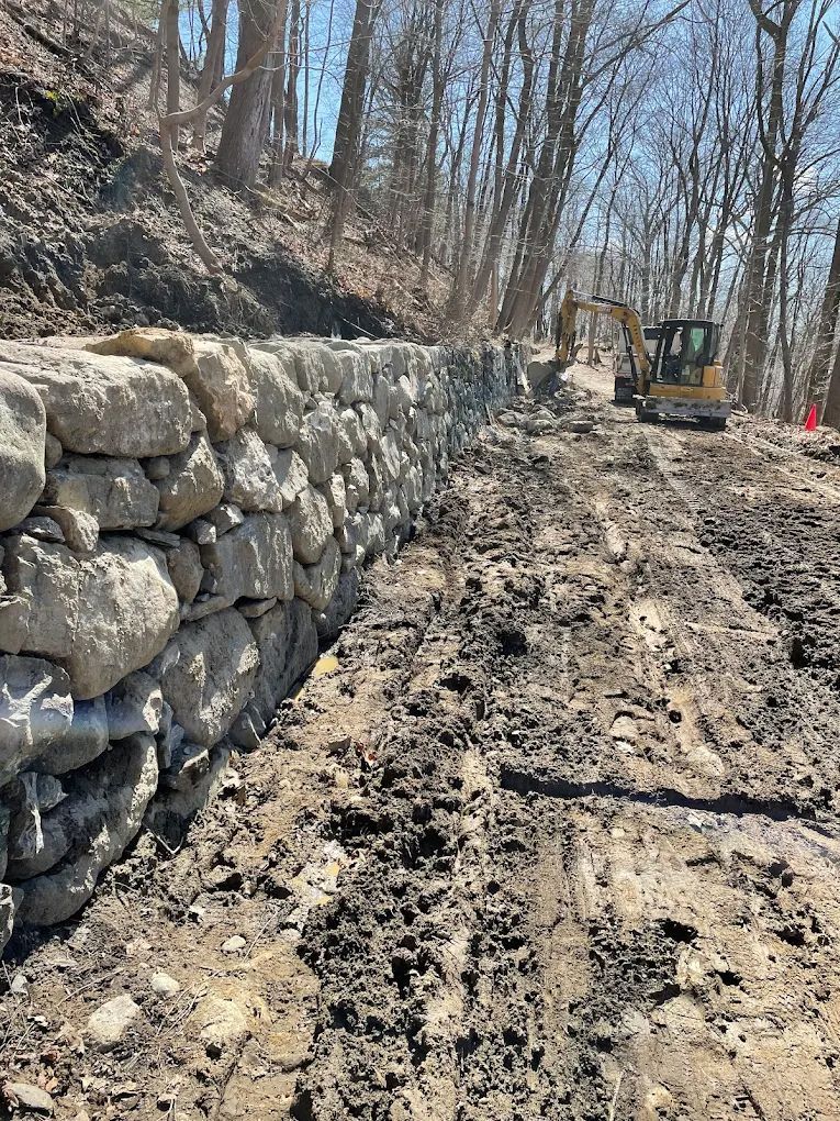Stone retaining wall under construction on a muddy road with an excavator.