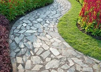 A curved stone pathway winds through a garden, flanked by green grass and colorful red foliage.