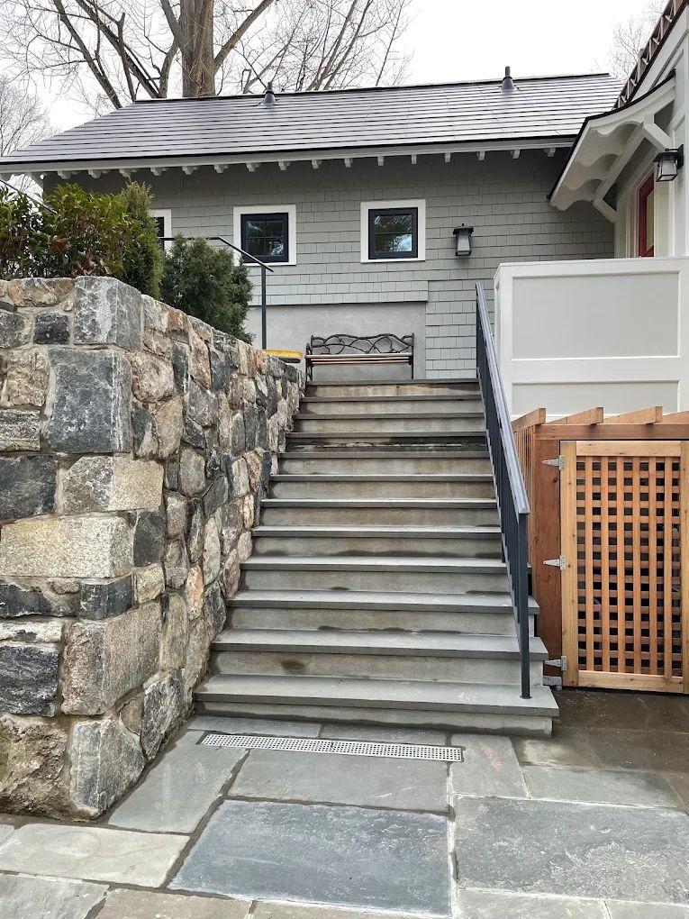 Stone stairs lead up to a gray building with two windows; a stone wall is to the left.