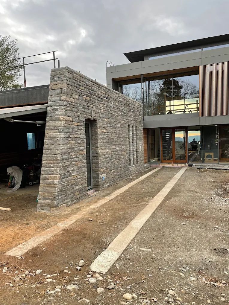 Stone wall and driveway lead to modern house with glass windows on a cloudy day.