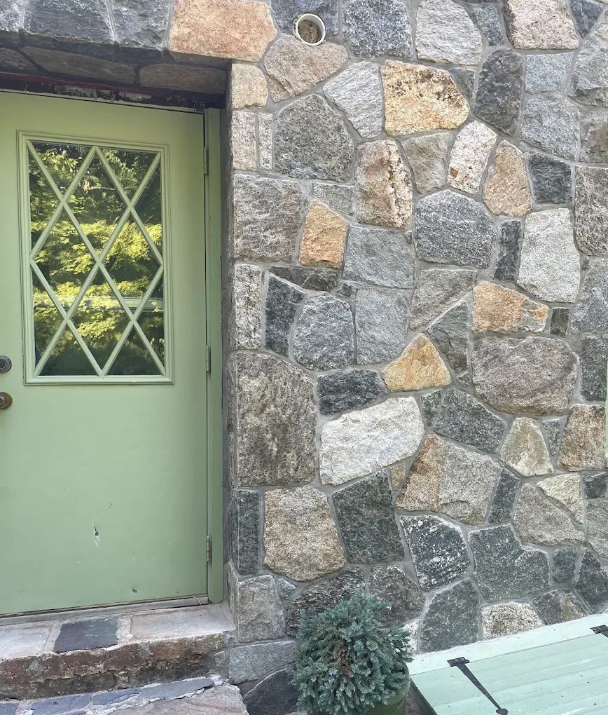 Green door with diamond-shaped window and a stone wall. Small bush in front.