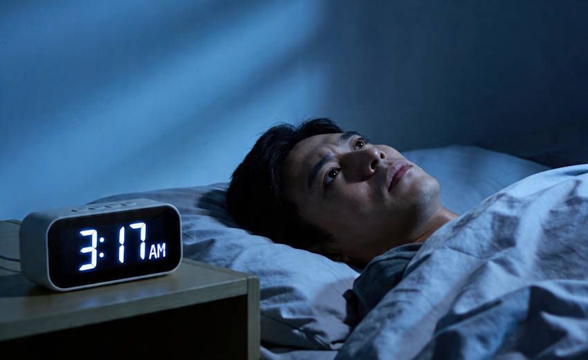 Asian man laying in bed staring at ceiling with his eyes open with his alarm clock reading 3:17 AM with a frustrated exhausted expression