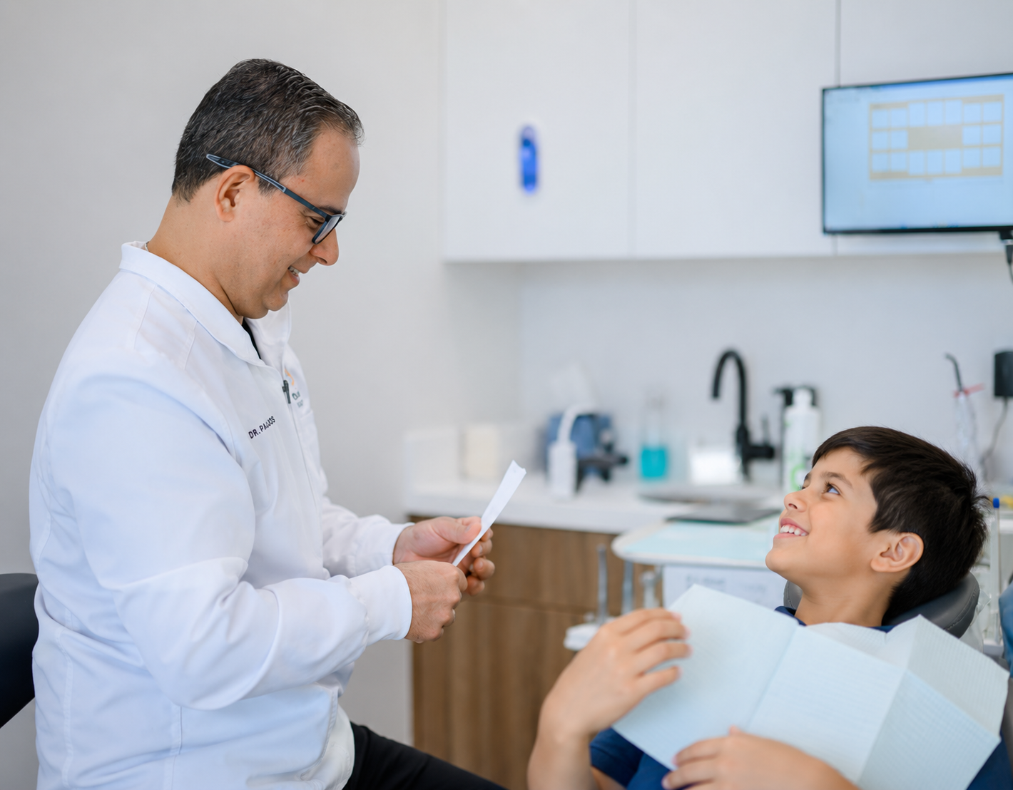 Dentist talking to a smiling patient in a modern clinic, holding an X-ray near a dental chair.
