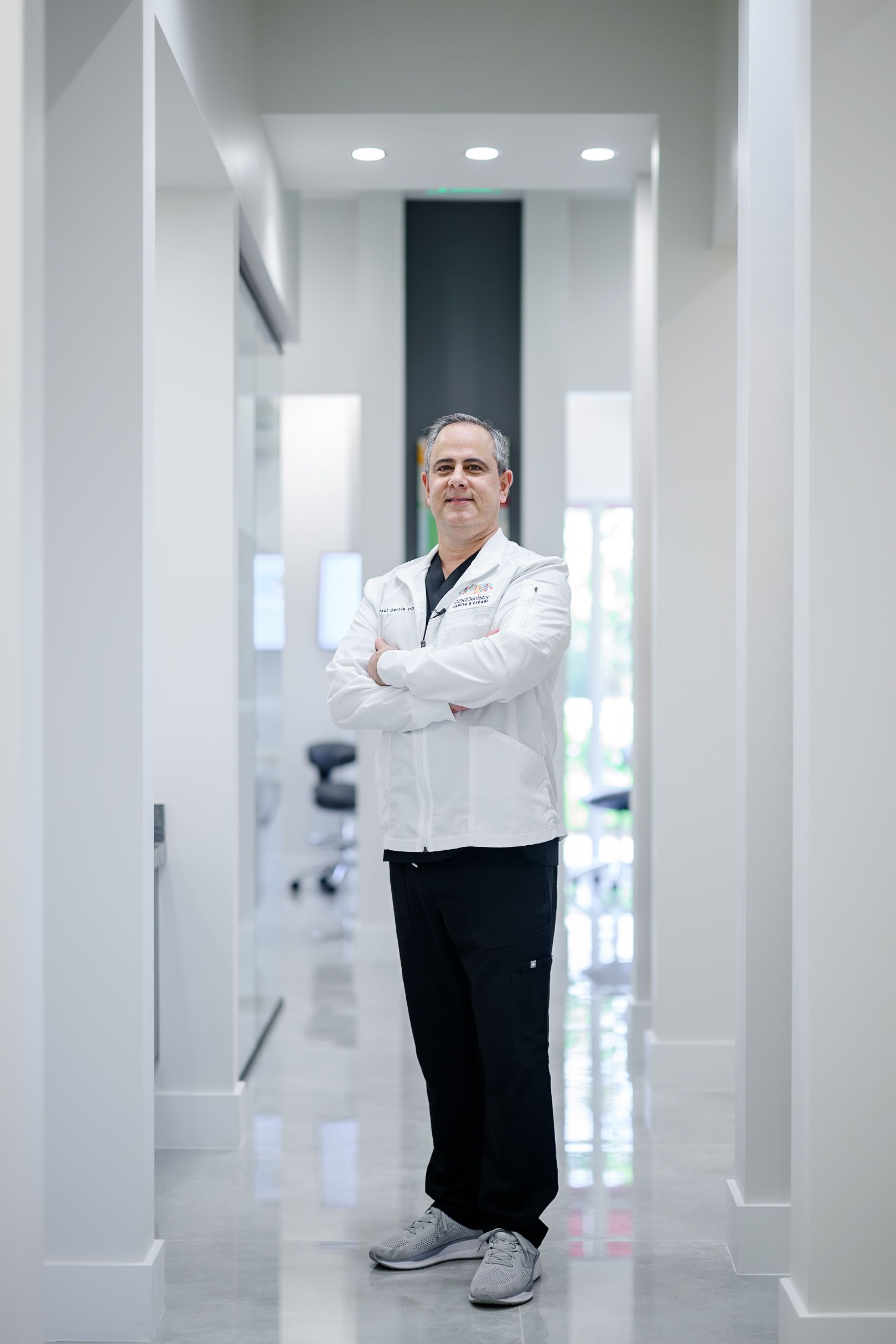 Person in a white lab coat standing in a bright modern hallway with arms crossed