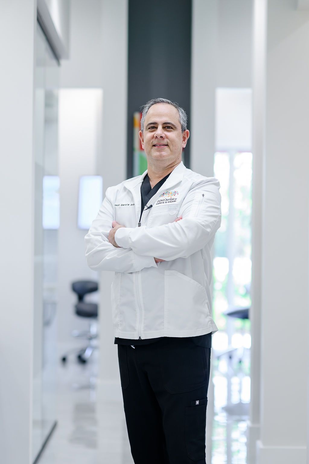 Doctor standing with arms crossed in a bright hospital hallway, wearing a white coat.