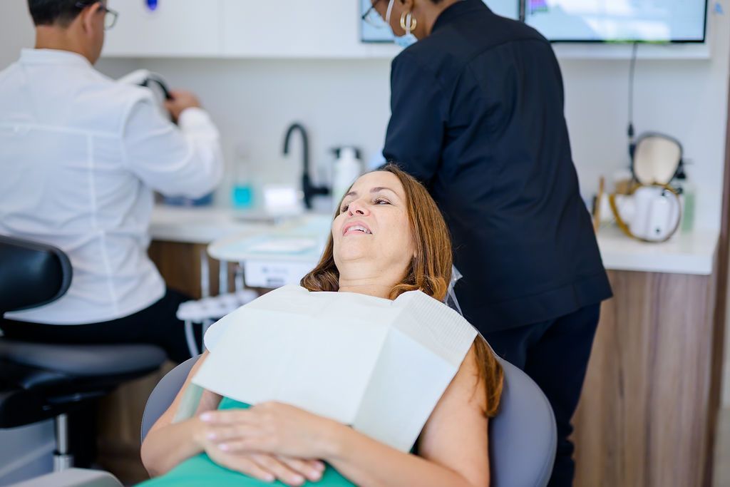 Woman reclines in a dental chair while two clinicians work in a bright dental office.