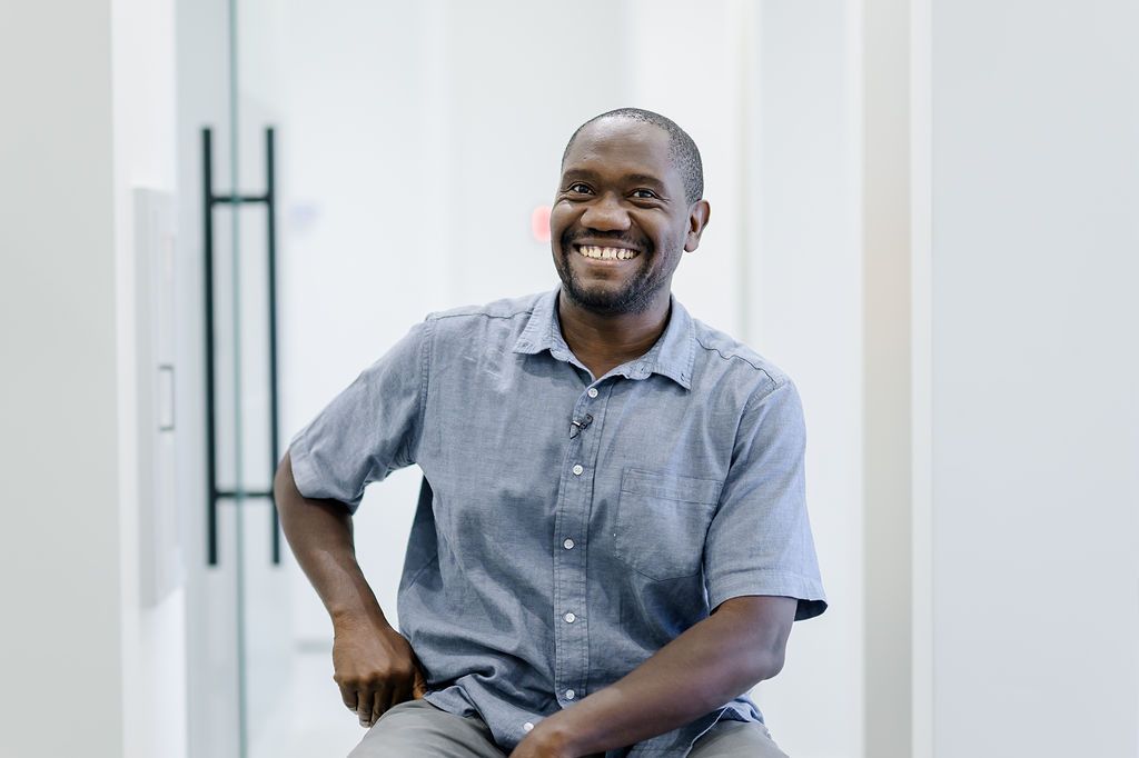 Smiling man seated indoors against a bright white background, wearing a gray button-up shirt.