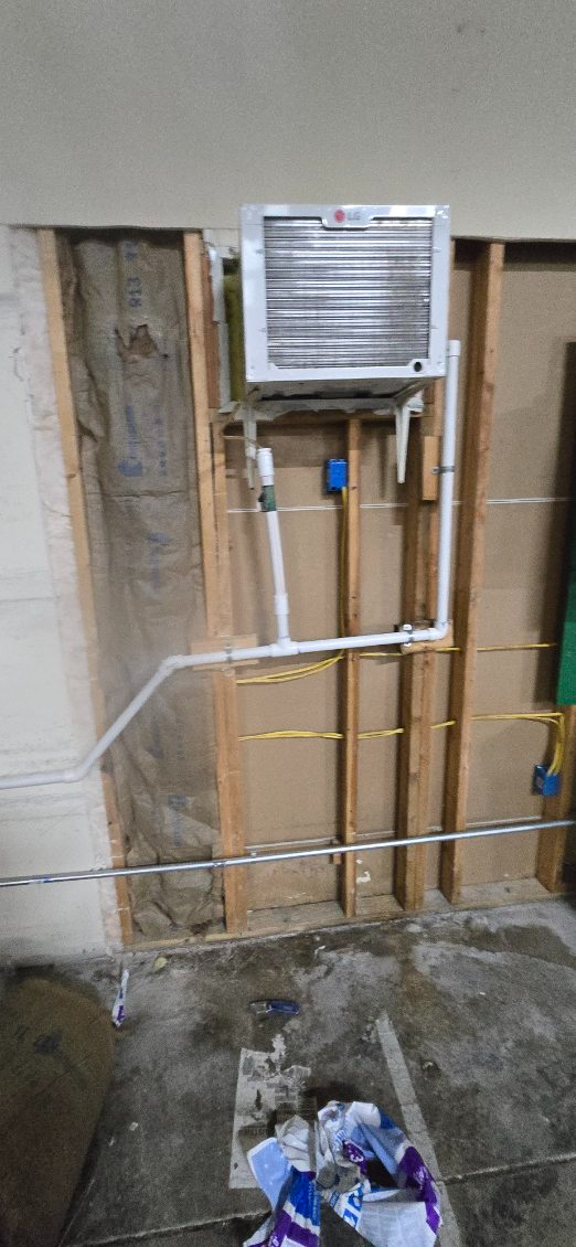 Wall with exposed framing, pipes, wiring, and an air conditioner unit. Concrete floor with debris.