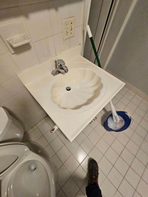 Small bathroom with a wall-mounted sink, tiled floor, toilet, and blue mop bucket; a shoe is visible at bottom.