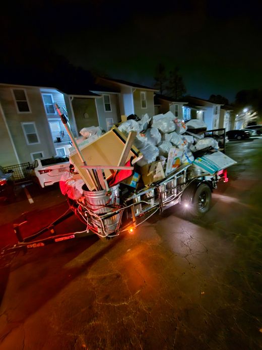Overflowing trailer loaded with debris and furniture on a wet street at night, with apartment buildings behind it