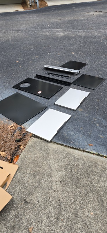 Various black and white metal sheets and parts arranged on asphalt pavement.