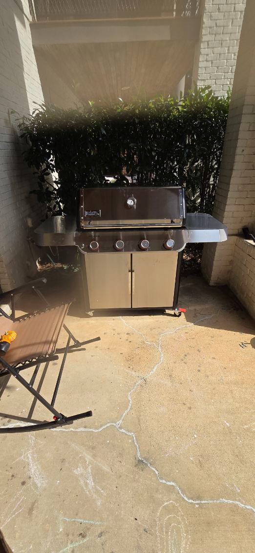 A stainless steel grill stands on a patio, backed by green hedges. A folding chair is nearby.