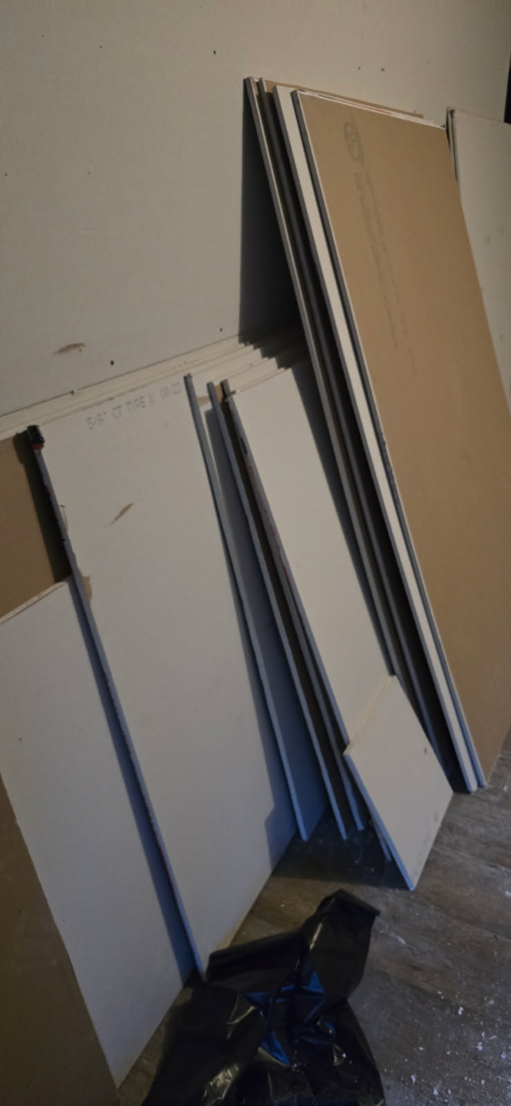 Unassembled furniture components stacked against a wall. Includes boards, metal supports and a black trash bag.