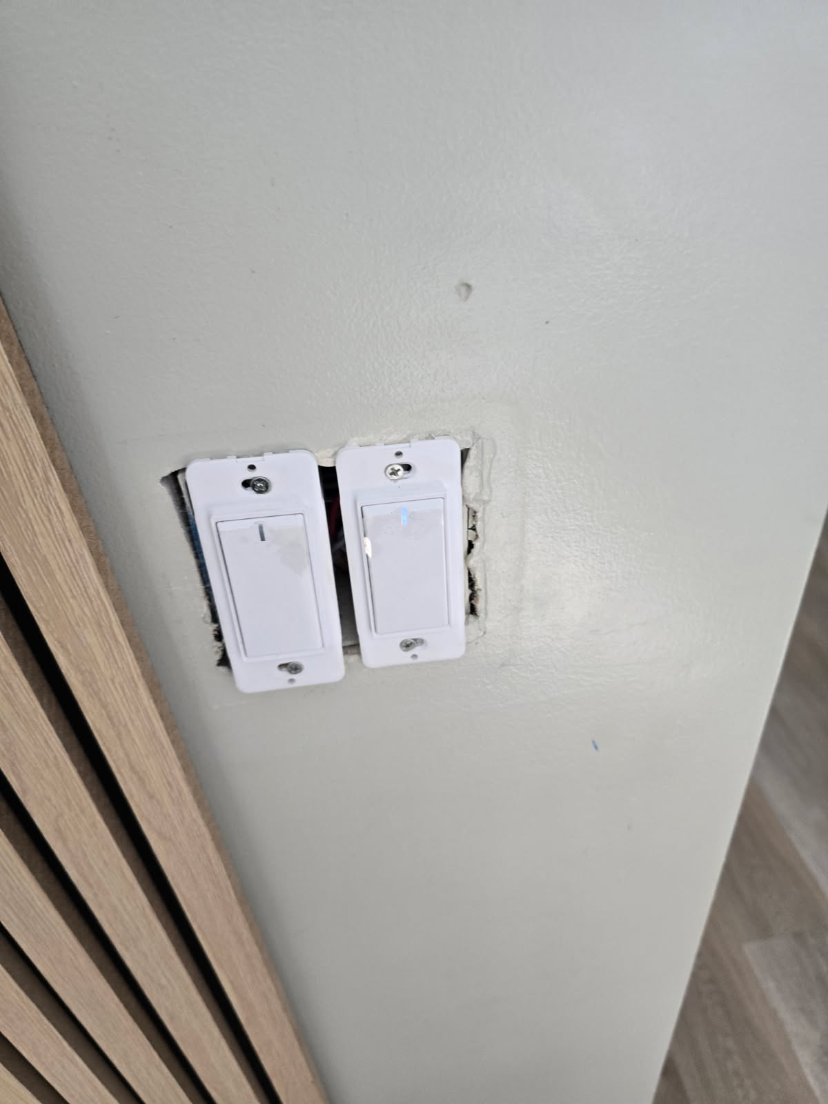 Two white light switches in a wall, slightly askew. Beige wall and wooden paneling in view.