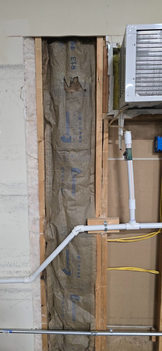 Wall with exposed insulation, framing, and PVC pipes. An air conditioning unit is in the background.