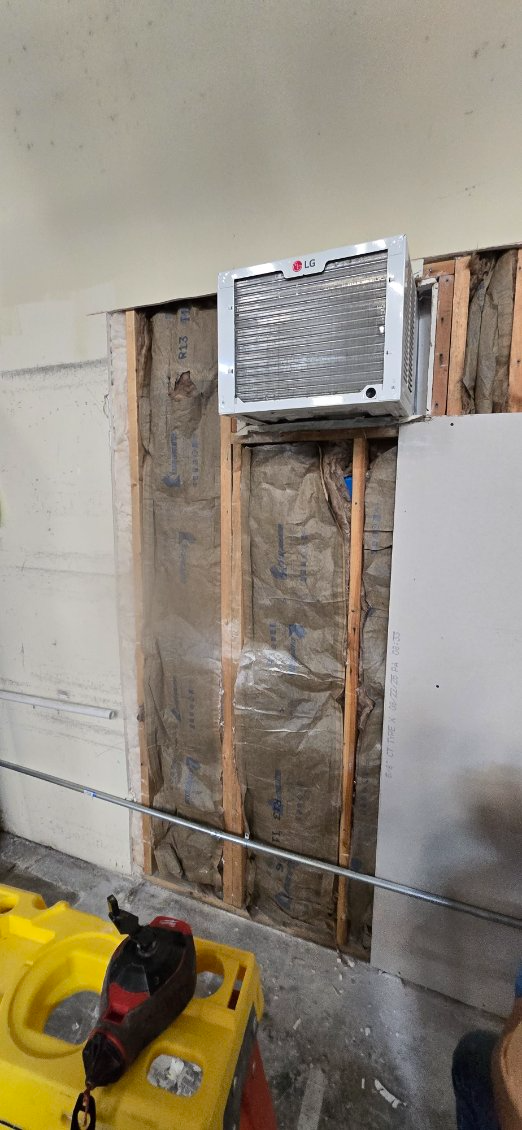 An air conditioner is installed in a wall with exposed framing. A jackhammer sits nearby.
