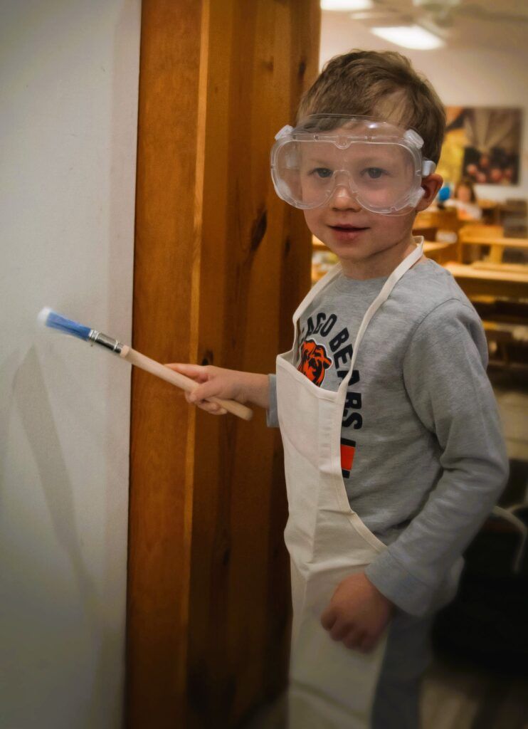 A young boy wearing goggles and an apron is holding a brush.