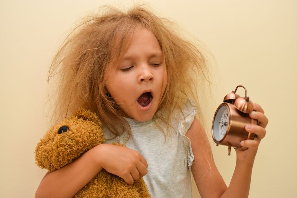 A little girl with messy hair is holding a teddy bear and holding an alarm clock.