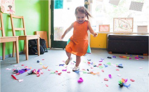 A little girl in an orange dress is playing with confetti on the floor.