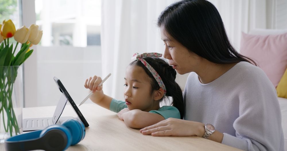 Woman helps child with tablet, indoors. Table with flowers, headphones.