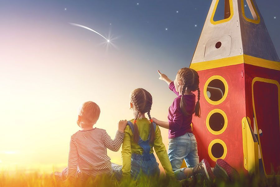 Three children are sitting in front of a cardboard rocket looking at the stars.