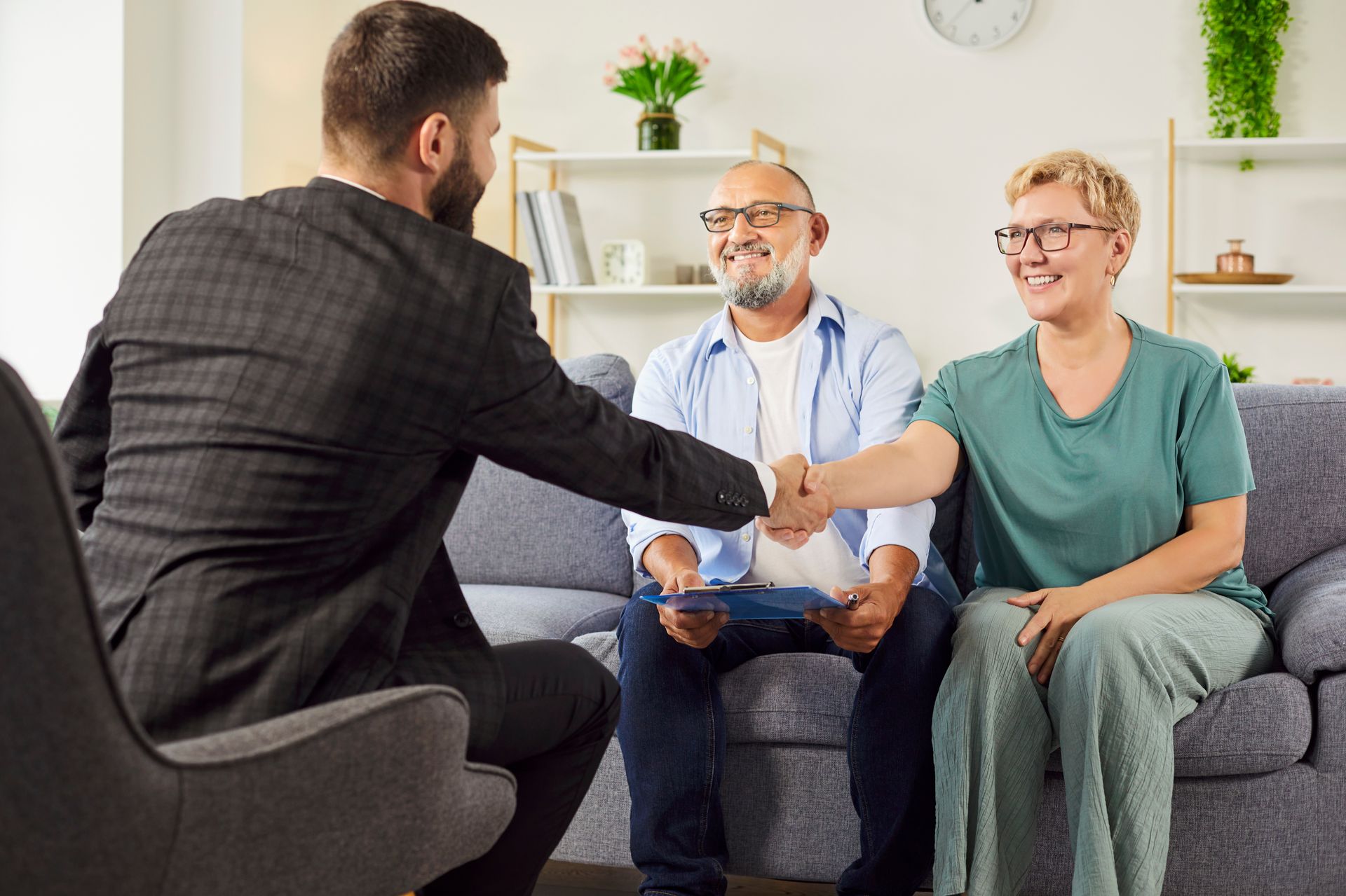 Man in suit shakes hands with a couple on a sofa. They are smiling in a home setting.