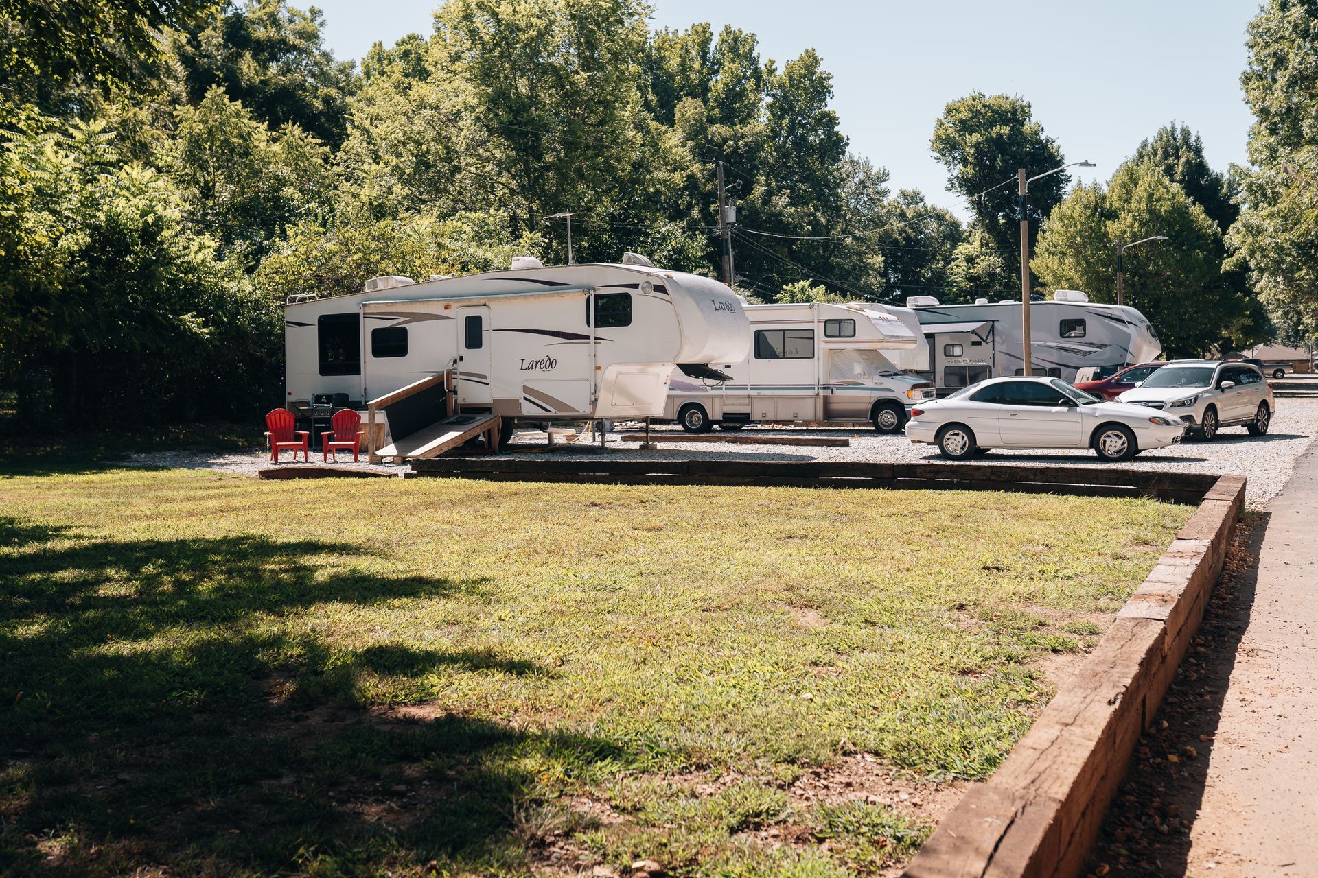 Several recreational vehicles and cars parked in a grassy campground shaded by tall, green trees.