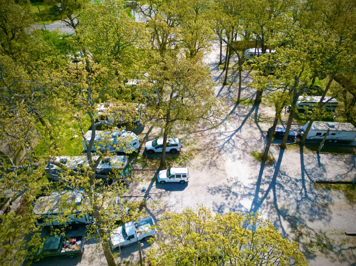 Aerial view of a wooded campground featuring several parked recreational vehicles and cars on a gravel lot.