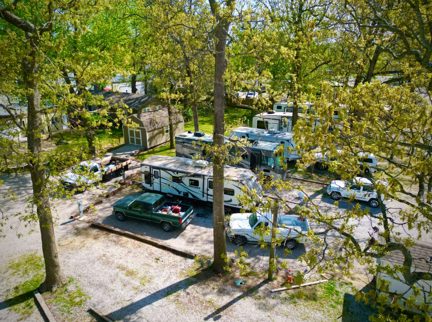 An aerial view of a wooded RV park with several campers and parked vehicles in a gravel lot.