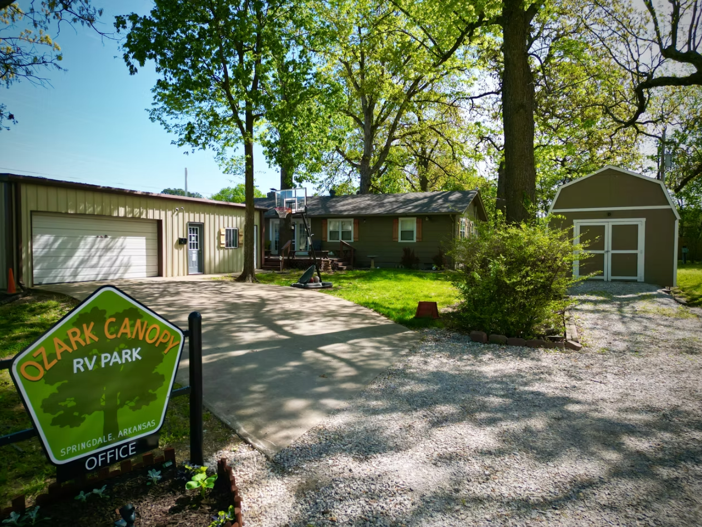 An outdoor scene showing the Ozark Canopy RV Park office, a house, and a shed on a gravel and concrete driveway.