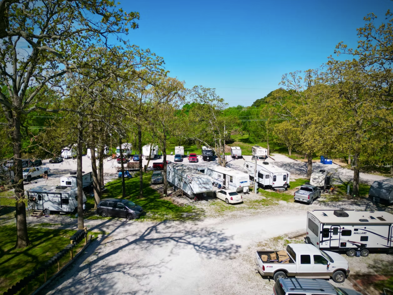 An aerial view of a wooded RV park with multiple campers, trailers, and parked vehicles on a gravel lot under a clear sky.