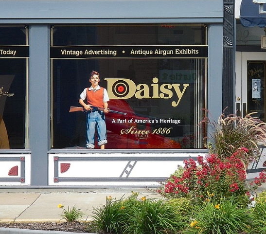 A Daisy museum storefront window displaying a cutout of a person with a toy rifle, advertising vintage airgun exhibits.